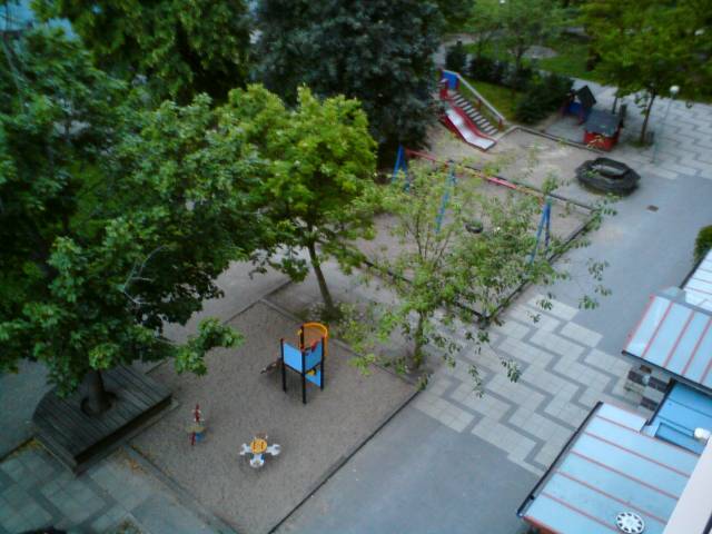Foto taget från min balkong. Den visar ett träd, lekplats i sand, stenlagd väg framför huset, glastak på uteplatser med mera. 
