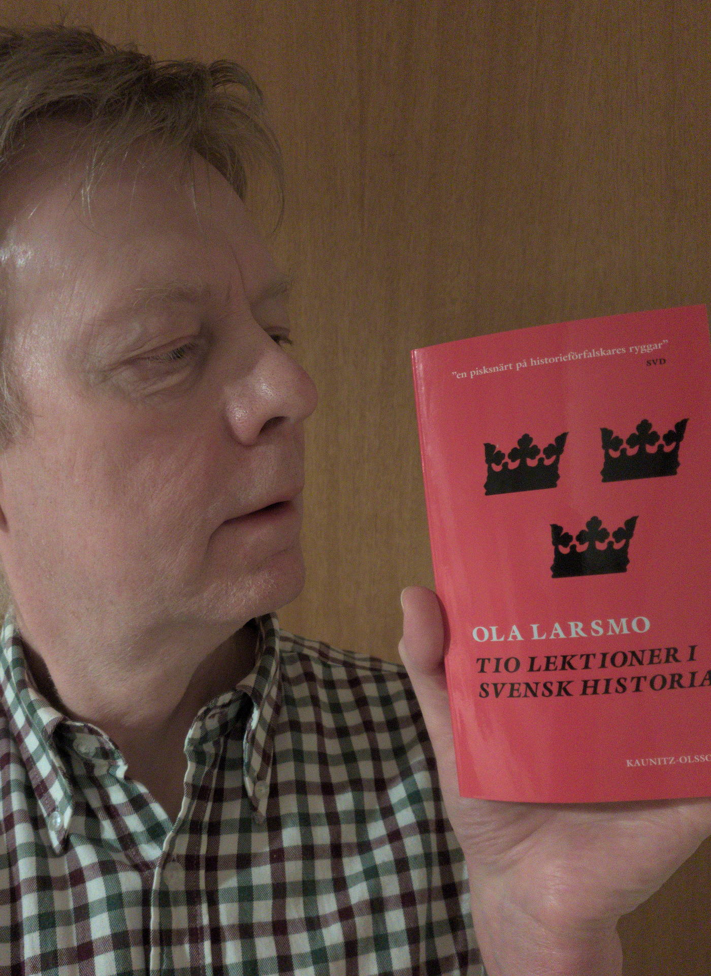 Jag (Henrik Hemrin) håller boken Tio lektioner i svenska historia av Ola Larsmo i min vänstra hand. Bokens omslag är i rött och jag har en smårutig skjorta.