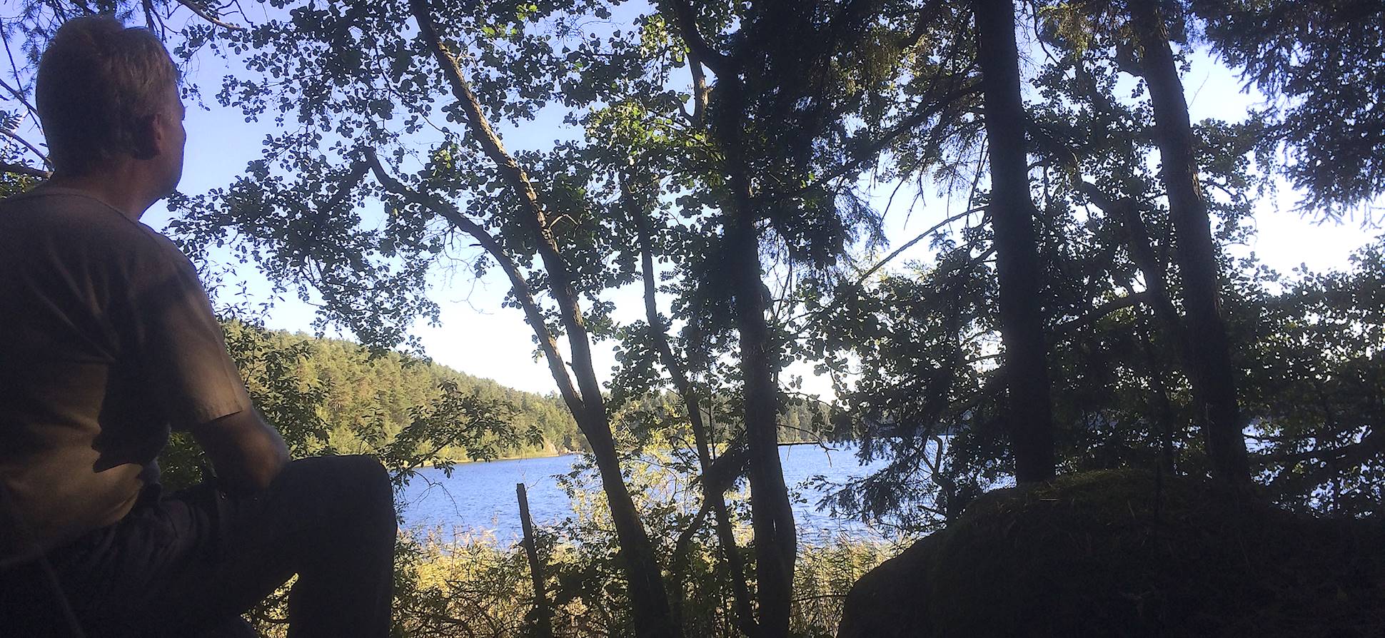 Self portrait from behind. I sit and look out over a lake, many trees both in foreground and at left of the lake. Blue sky with some clouds. Photo: Henrik Hemrin. 